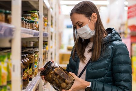 Woman in medical mask holding glass jar of pickled food in a supermarket.