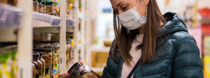 Woman in medical mask holding glass jar of pickled food in a supermarket.